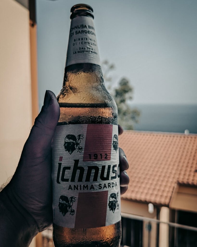 Hand holding Ichnusa beer bottle with Sardinian background.