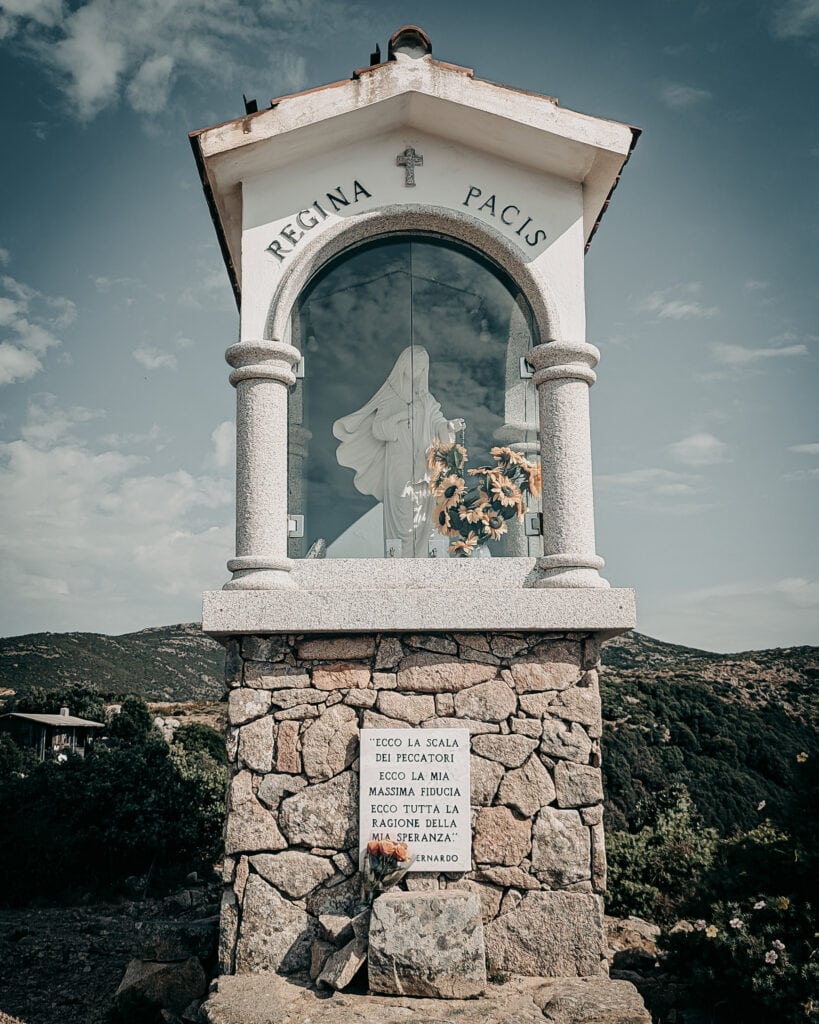 Small religious shrine with statue and flowers in a scenic landscape.