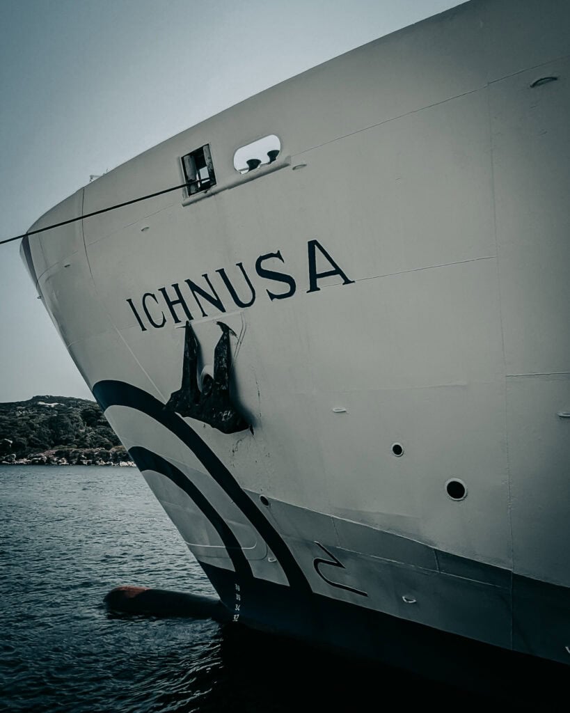 Close-up of the cruise ship Ichnusa docked at port with water and landscape in background.