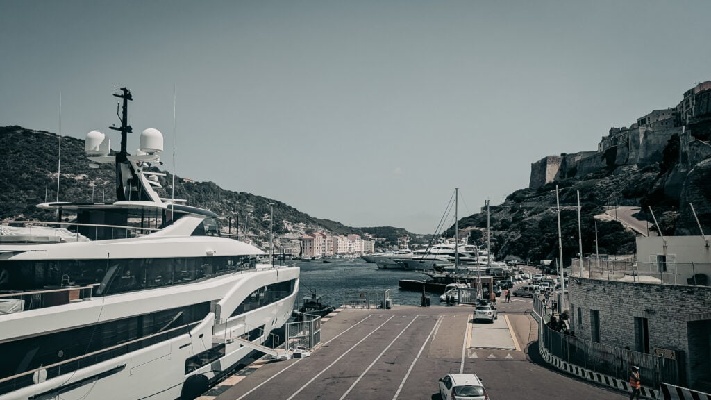 Luxury yacht moored at a Mediterranean harbor with hillside town in background.